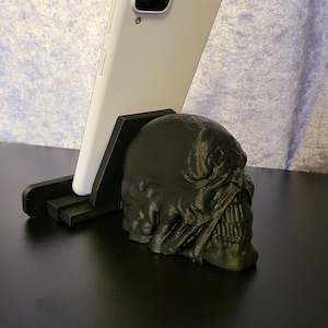 May include: A black skull-shaped phone stand supporting a white smartphone. The stand is designed to hold a phone upright. The skull has detailed features and is made of a dark material. The phone is white with a camera.