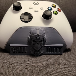 May include: A white Xbox controller sits on a grey stand with a skull design and the words "CALL DUTY". The controller has black joysticks and coloured buttons. The stand is 3D printed.
