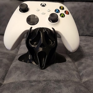 May include: A white Xbox controller sits on a black 3D-printed stand, designed as a Scream mask. The controller has black buttons and coloured action buttons. The stand is on a grey surface.