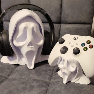 May include: White Ghostface-themed headphone and controller stands. The headphone stand features the iconic Ghostface mask with a hood, holding a pair of black headphones. A matching controller stand holds a white Xbox controller. Both are on a grey surface.