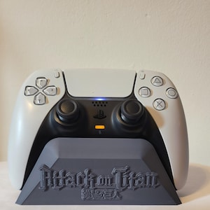 May include: A white and black wireless game controller resting on a dark grey plastic charging stand. The stand features the embossed text Attack on Titan in English and Japanese characters. The controller has two black analogue sticks.
