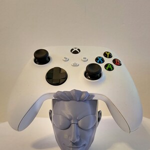May include: A white Xbox controller sits on a grey, 3D-printed head-shaped stand. The controller has black joysticks and coloured buttons. The stand features a detailed face with glasses and hair, designed to hold the controller.