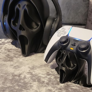 May include: Black Ghostface mask-shaped stands for a headset and a PlayStation 5 controller. The headset stand holds a black and grey headset. The controller stand supports a white and black controller.