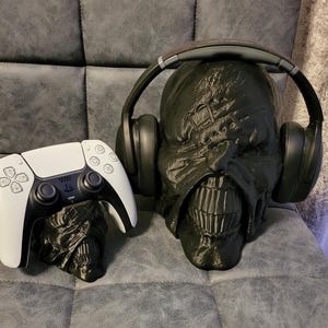 May include: Black 3D-printed Nemesis head with headphones and a white and black PlayStation 5 controller. The Nemesis head is a gaming accessory, designed to hold headphones. The controller rests on a smaller Nemesis head.