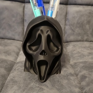 May include: A black Ghostface mask pen holder, inspired by the Scream film series. The pen holder is made of a dark material and holds pens in blue, teal, and silver. The pen holder is on a grey surface. The pen holder is approximately 15 cm tall.