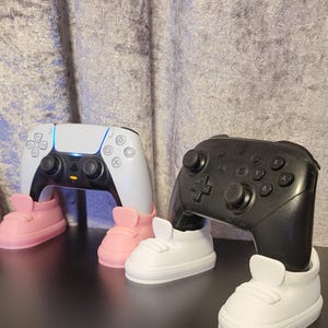 May include: A white and black PlayStation 5 controller and a black Nintendo Switch Pro controller, each supported by a pair of miniature trainers. The trainers are in pink and white. The controllers are on a black surface.
