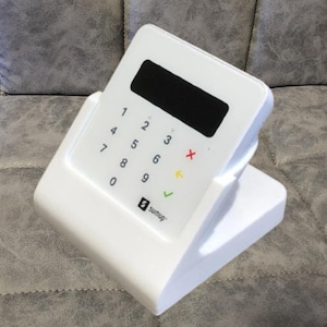 May include: A white SumUp card reader with a black display screen and a numeric keypad. The keypad includes numbers, a red X, a yellow arrow, and a green tick. The device is on a white stand.