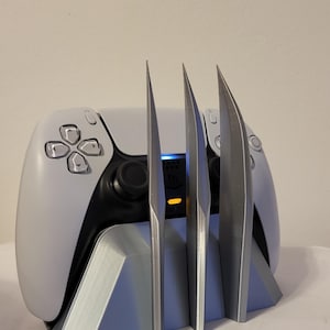 May include: A white and black video game controller resting in a silver-coloured stand. The stand features three vertical, pointed, blade-like protrusions extending upwards in front of the controller, which has a glowing blue and orange light.