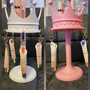 May include: Two crown-shaped cosmetic organisers, one white and one pink, each with a central post and a circular base. Small tubes of skincare products hang from the crown's lower edge. The organisers hold pink and gold cosmetic tubes.