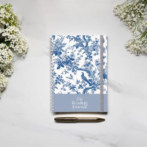 May include: A blue and white spiral bound journal with a floral pattern and the text "The Reading Journal" on the cover. A gold pen is lying next to the journal.