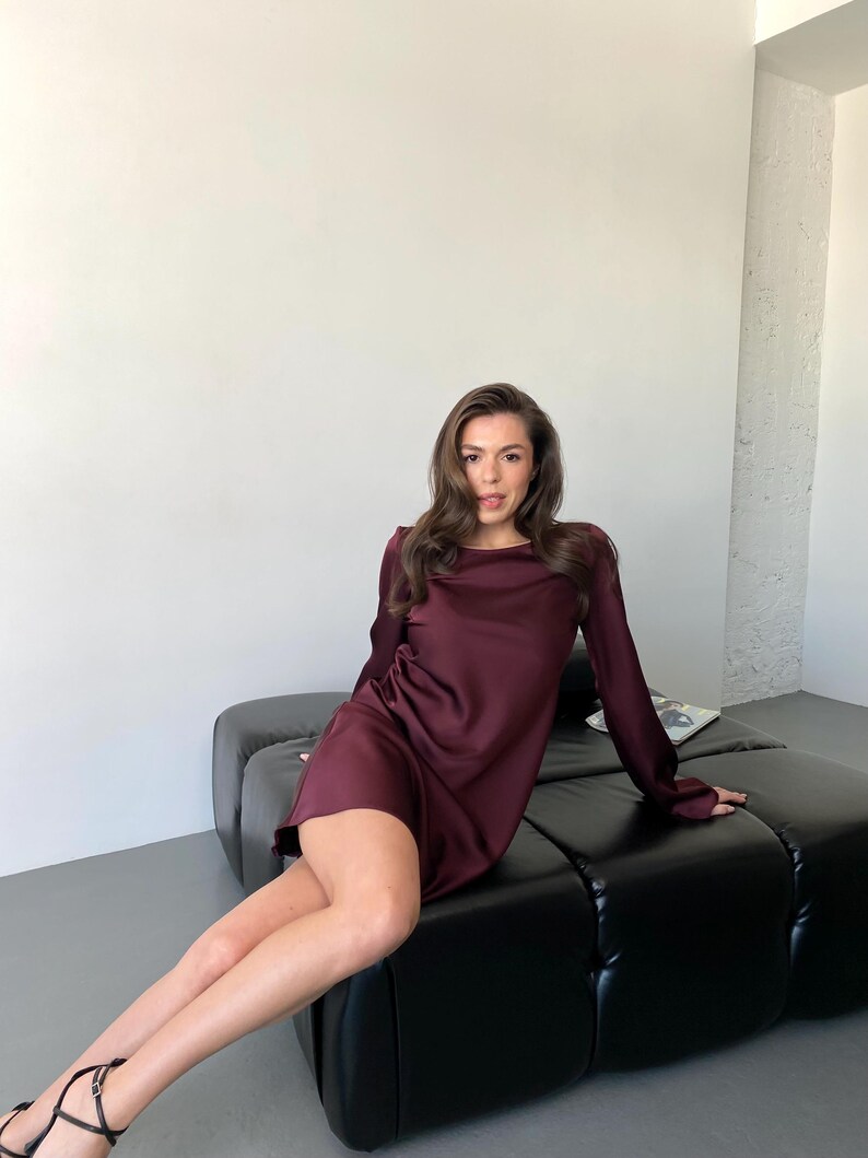 May include: A burgundy-colored, long-sleeve dress with a draped neckline. The dress is made of a silky material and is knee-length. The model is sitting on a black leather couch.