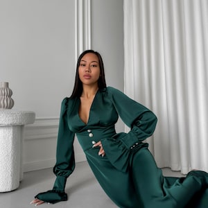 May include: A woman wearing a long, emerald green satin dress with a deep V-neckline and pearl buttons. The dress has long sleeves with ruffles at the cuffs. The woman is posing on a light gray floor.