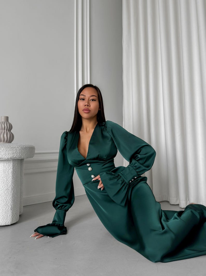 May include: A woman wearing a long, emerald green satin dress with a deep V-neckline and pearl buttons. The dress has long sleeves with ruffles at the cuffs. The woman is posing on a light gray floor.