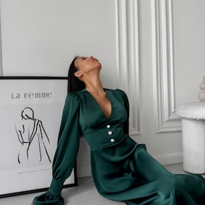 May include: A woman wearing a deep green satin dress with long sleeves and a V-neckline. The dress has pearl buttons at the waist. The woman is sitting on the floor with her head tilted back. The background is a white wall with a framed print that says "La femme" and a drawing of a female figure.