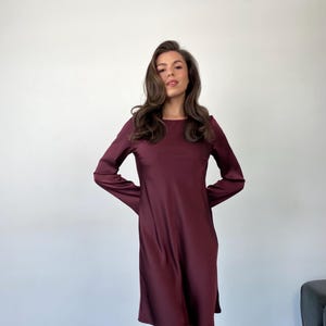 May include: A burgundy-colored long-sleeve dress with a round neckline. The dress has a simple, flowing design and falls to about knee length. The fabric appears to be smooth and has a slight sheen.