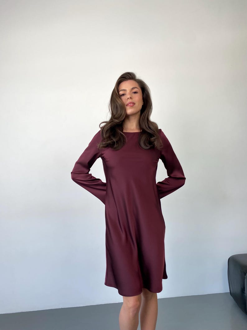 May include: A burgundy-colored long-sleeve dress with a round neckline. The dress has a simple, flowing design and falls to about knee length. The fabric appears to be smooth and has a slight sheen.