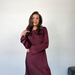 May include: A burgundy-colored dress with long sleeves and a round neckline. The dress has a simple, elegant design and falls to about knee length. The fabric appears to be a smooth, satin-like material. The dress is suitable for various occasions.