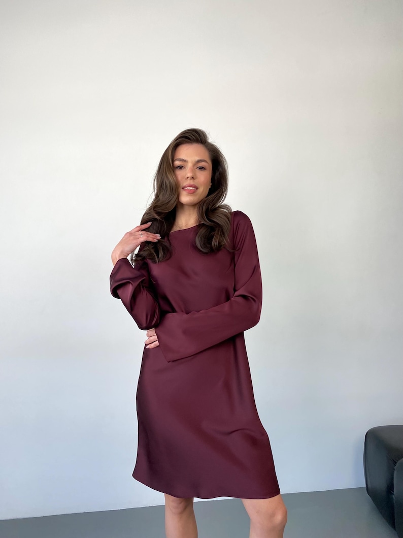 May include: A burgundy-colored dress with long sleeves and a round neckline. The dress has a simple, elegant design and falls to about knee length. The fabric appears to be a smooth, satin-like material. The dress is suitable for various occasions.
