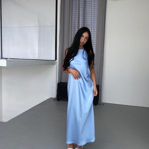 Puede incluir: Un vestido largo de satén azul claro con cuello halter. El vestido se lleva con sandalias blancas. La modelo tiene el pelo largo y oscuro.