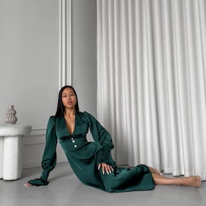 May include: A woman wearing a long-sleeved, emerald green satin dress with a deep V-neckline and pearl buttons. The dress has a fitted bodice and a flowing skirt. She is sitting on a light gray floor with a white curtain in the background.