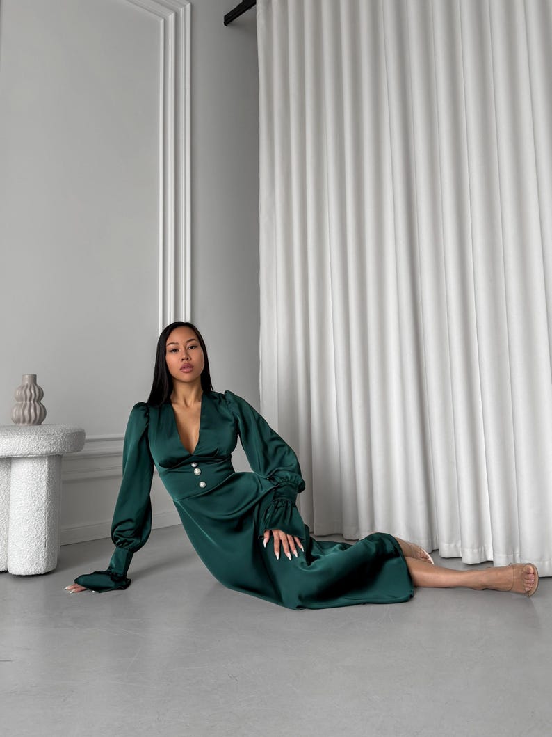 May include: A woman wearing a long-sleeved, emerald green satin dress with a deep V-neckline and pearl buttons. The dress has a fitted bodice and a flowing skirt. She is sitting on a light gray floor with a white curtain in the background.