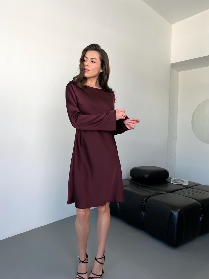 May include: A burgundy-colored dress with long sleeves and a flared skirt. The dress has a boat neckline and is knee-length. The model is wearing black strappy heels. The background is a minimalist interior with a black leather ottoman.