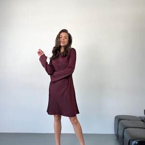 May include: A burgundy-colored dress with long sleeves and a round neckline. The dress falls to the mid-thigh and is paired with black strappy heels. The model is standing in front of a white wall.