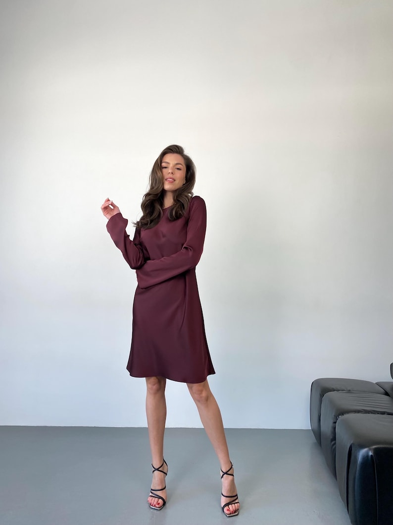 May include: A burgundy-colored dress with long sleeves and a round neckline. The dress falls to the mid-thigh and is paired with black strappy heels. The model is standing in front of a white wall.