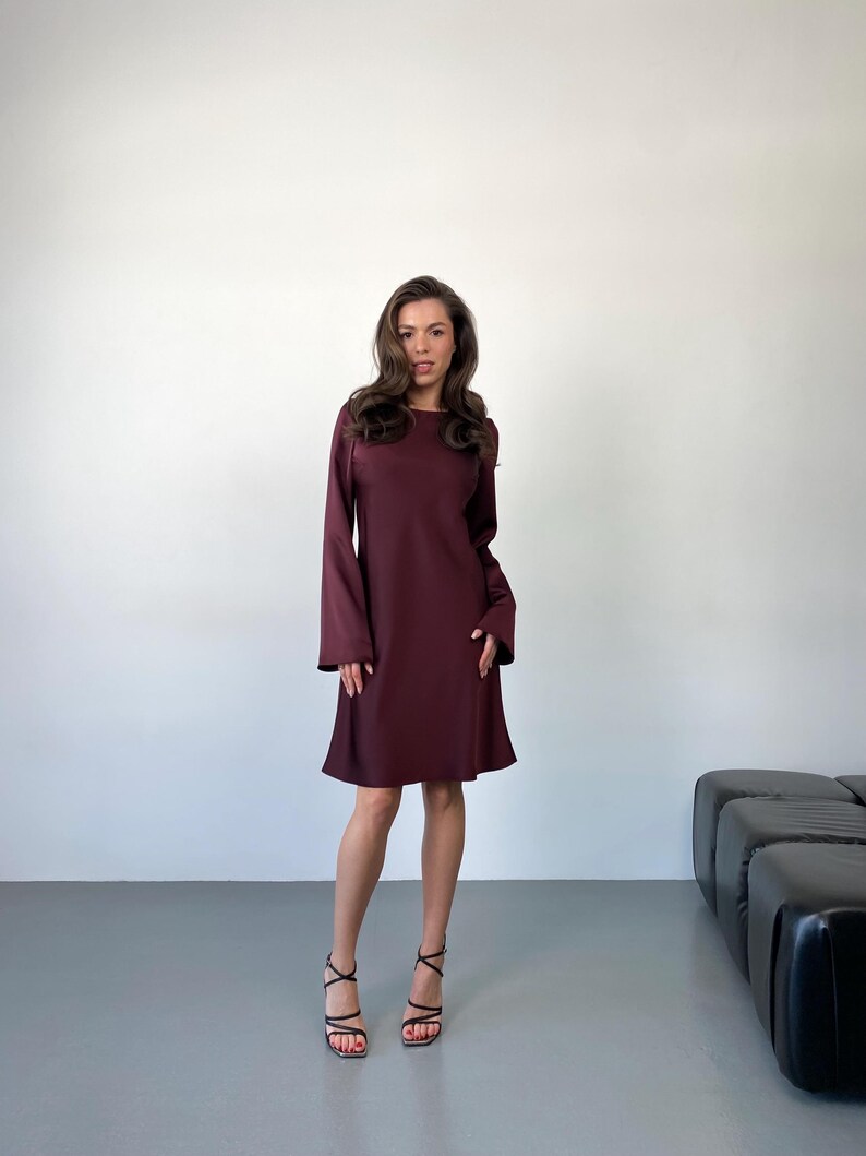 May include: A maroon-colored dress with long sleeves and a flared skirt. The dress is knee-length and has a simple, elegant design. The model is wearing black strappy heels.