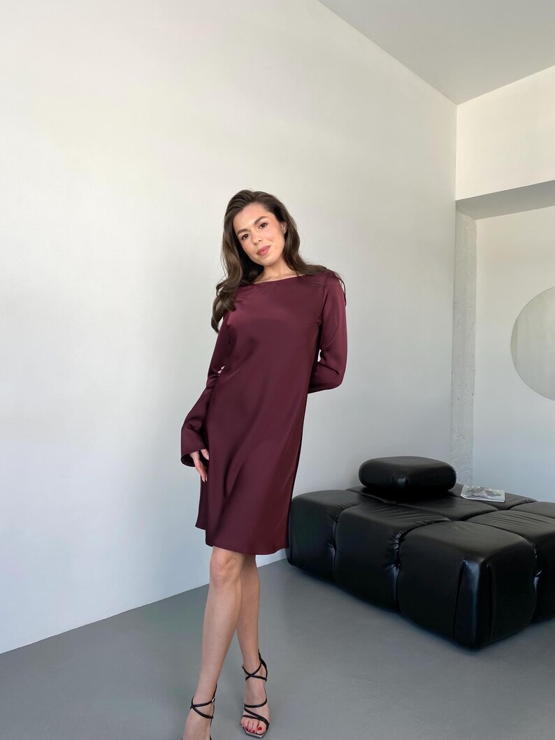 May include: A long-sleeved, knee-length dress in a deep burgundy color. The dress has a simple, elegant design with a round neckline and a slightly loose fit. The model is wearing black strappy heels.