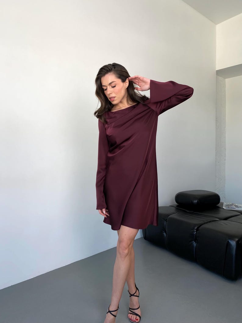 May include: A burgundy-colored, long-sleeve dress with a draped neckline. The dress has a flowing, satin-like texture and falls to the mid-thigh. The model is wearing black high-heeled sandals. The background is a plain white wall and a black leather couch.