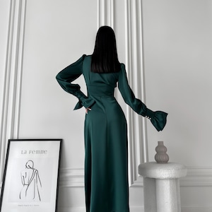 May include: A woman wearing a long, dark green satin dress with a V-neckline and long sleeves. The dress has a fitted bodice and a flowing skirt. The woman is standing in front of a white wall with a framed print that says "La Femme".