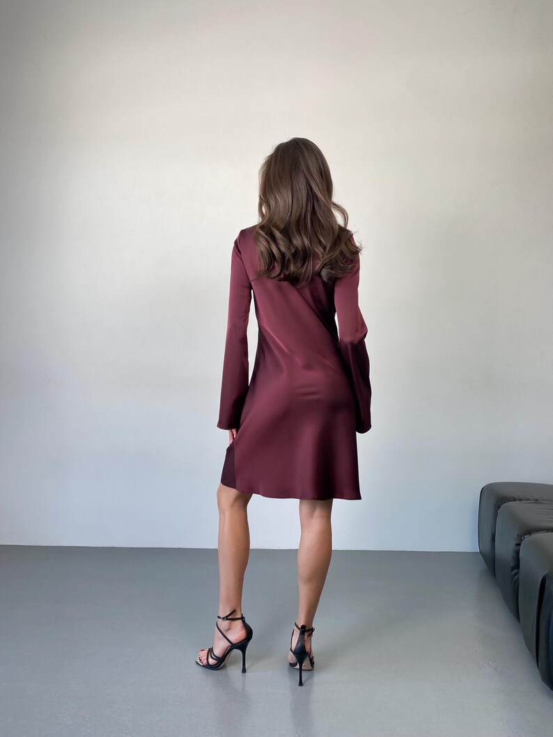 May include: A burgundy-colored dress with long sleeves and a flared skirt. The dress is paired with black high-heeled sandals. The model is standing in front of a plain white wall and a gray floor.