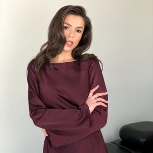 May include: A long-sleeved, burgundy dress with a boat neckline. The dress is made of a smooth, satin-like material. The model is wearing the dress with her arms crossed.