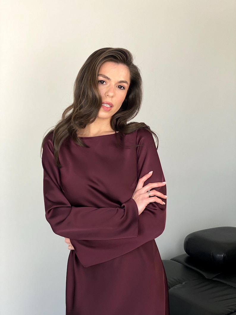 May include: A long-sleeved, burgundy dress with a boat neckline. The dress is made of a smooth, satin-like material. The model is wearing the dress with her arms crossed.
