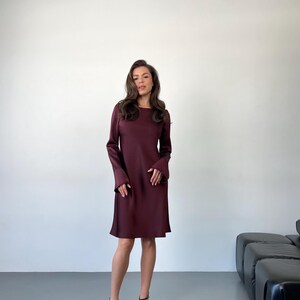 May include: A burgundy-colored dress with long sleeves and a knee-length hemline. The dress has a simple, elegant design. The person is wearing black strappy heels.