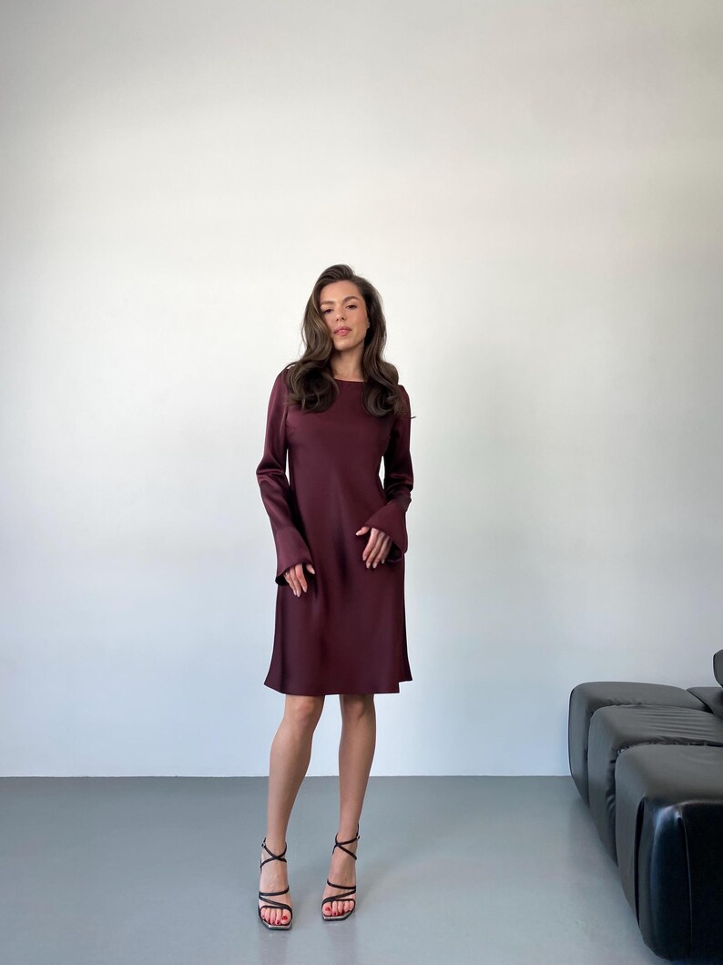 May include: A burgundy-colored dress with long sleeves and a knee-length hemline. The dress has a simple, elegant design. The person is wearing black strappy heels.