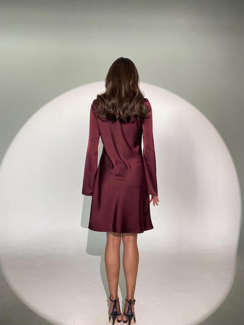 May include: A burgundy-colored dress with long sleeves and a flared skirt. The dress is made of a smooth, shiny fabric. The model is wearing black high-heeled sandals. The background is a white studio backdrop with a bright spotlight.