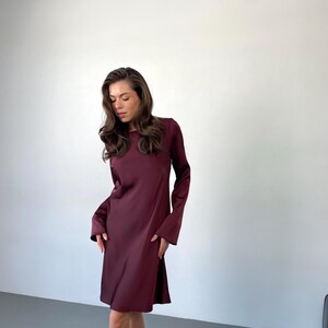 May include: A burgundy-colored dress with long sleeves and a flared skirt. The dress has a round neckline and a simple, elegant design. The fabric appears to be a smooth, flowing material. The dress is knee-length and suitable for various occasions.
