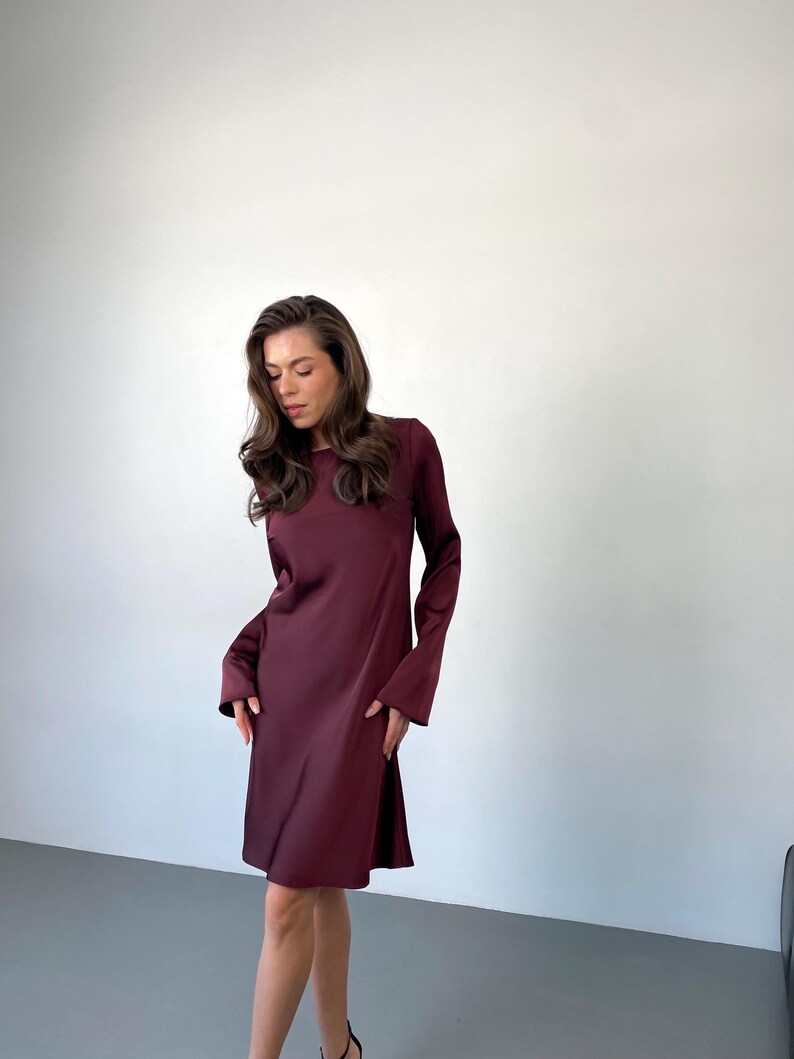 May include: A burgundy-colored dress with long sleeves and a flared skirt. The dress has a round neckline and a simple, elegant design. The fabric appears to be a smooth, flowing material. The dress is knee-length and suitable for various occasions.