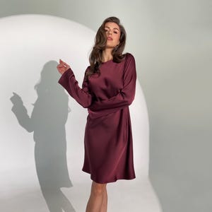 May include: A burgundy-colored, long-sleeve dress with a round neckline. The dress has a slightly flared skirt and is made of a smooth, silky material. The model is wearing black strappy heels. The background is a bright white.