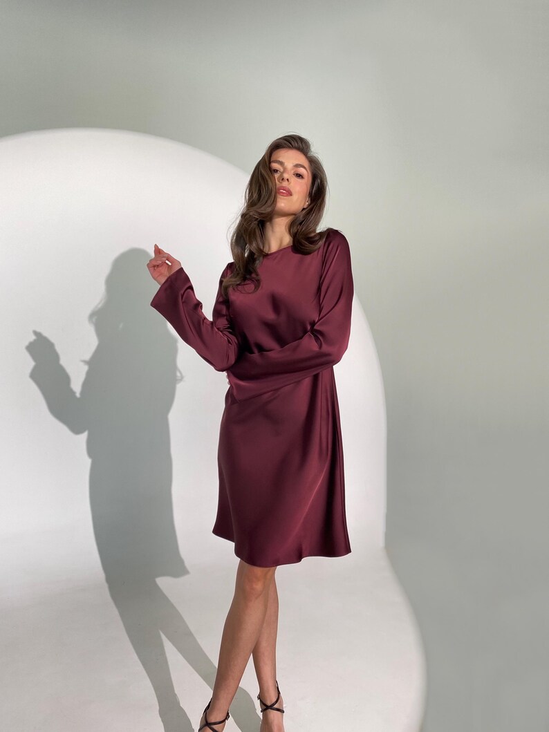 May include: A burgundy-colored, long-sleeve dress with a round neckline. The dress has a slightly flared skirt and is made of a smooth, silky material. The model is wearing black strappy heels. The background is a bright white.