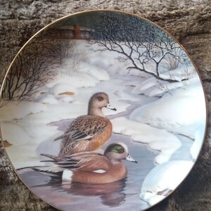 May include: A decorative porcelain plate featuring a winter scene with two American wigeon ducks in a snowy landscape. The plate has a gold rim and a signature at the bottom.
