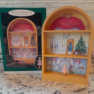 Peut inclure: Ornement d'étagère en bois jaune représentant une scène miniature du « Casse-Noisette ». L'ornement comporte deux étagères avec des décors peints d'une scène et d'un sapin de Noël. Une petite figurine de ballerine est devant l'étagère.