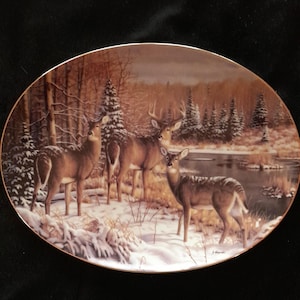 May include: A ceramic oval platter with a winter scene featuring three white-tailed deer in a snowy forest. The deer are standing in the snow near a small stream. The background is a forest with snow-covered trees.