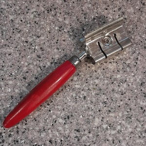 May include: A red handled metal pastry cutter with a silver blade. The cutter has a series of ridges on the blade.