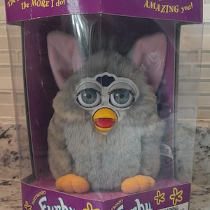 May include: A gray Furby toy in its original packaging. The electronic toy has large blue eyes, pink ears, and orange feet. The box is purple and has text that reads "The more you play with me the MORE I do!" and "I will KEEP AMAZING you!"