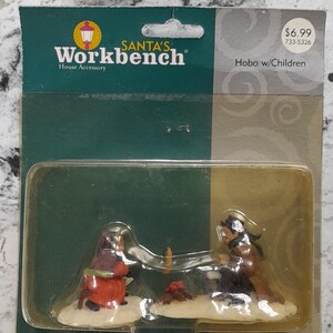 May include: A Christmas-themed figurine set from Santa's Workbench, featuring two figures near a small campfire. One figure wears a red coat, the other a brown coat. The package reads "Hobo w/Children."