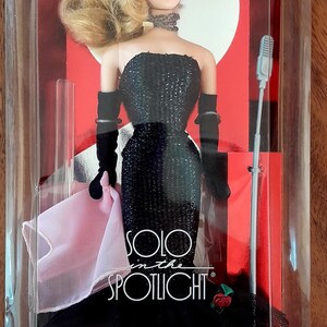 May include: A vintage Barbie doll in a clear plastic case. The doll wears a black sequined strapless gown, long black gloves, and a choker. The box reads "Solo in the Spotlight" and "Original 1960 Fashion & Doll!"