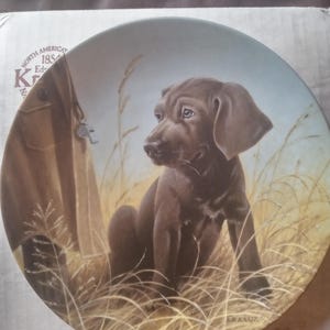 May include: A brown and white ceramic plate with a painting of a brown puppy sitting in tall grass. The plate has a gold rim and the words "North American 1854 Ed. Knowles" in a gold script font.